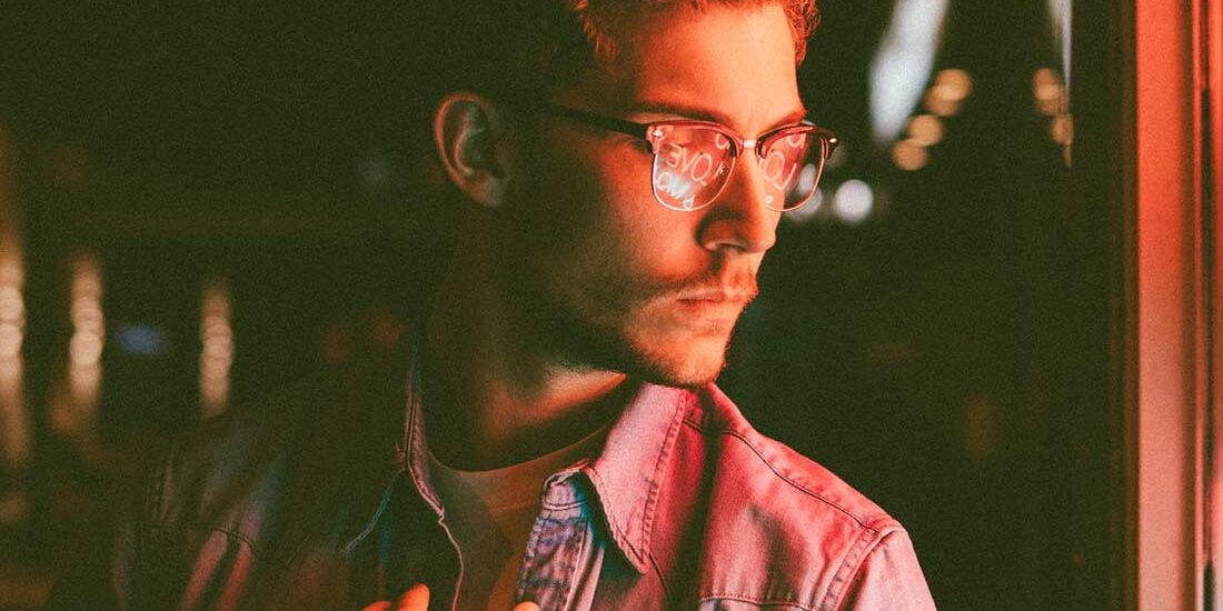 A man with glasses standing by a window with warm lighting.