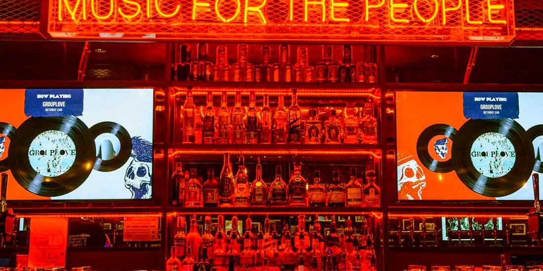 A neon sign reading 'MUSIC FOR THE PEOPLE' above a bar with two screens showing album art.