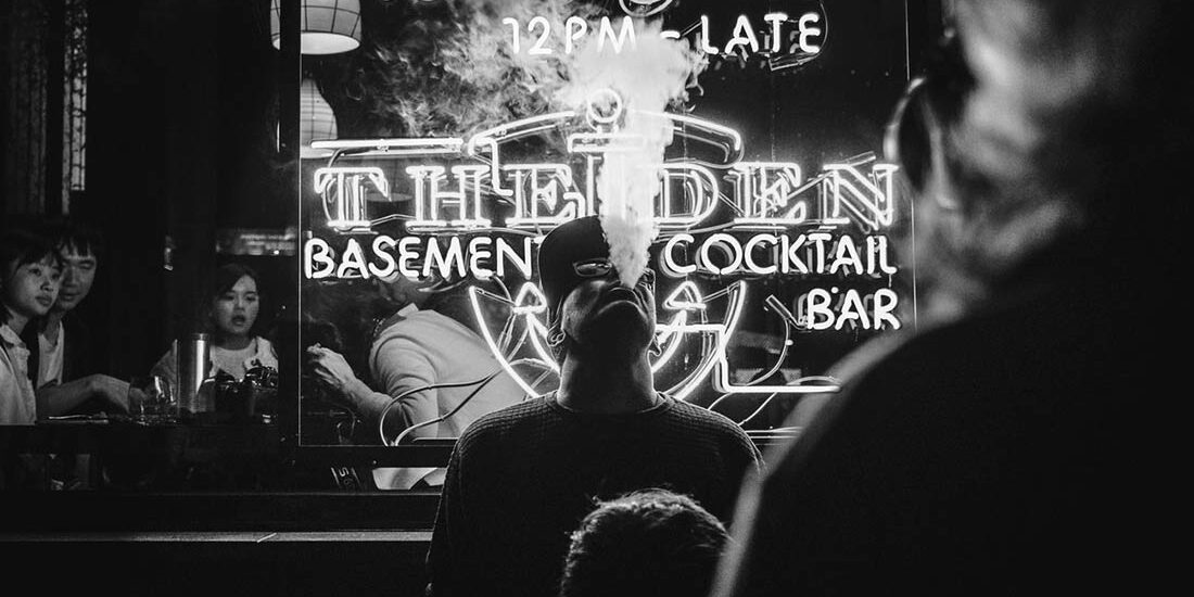 Black and white photo of a jazz musician playing trumpet in front of a neon-lit bar sign.