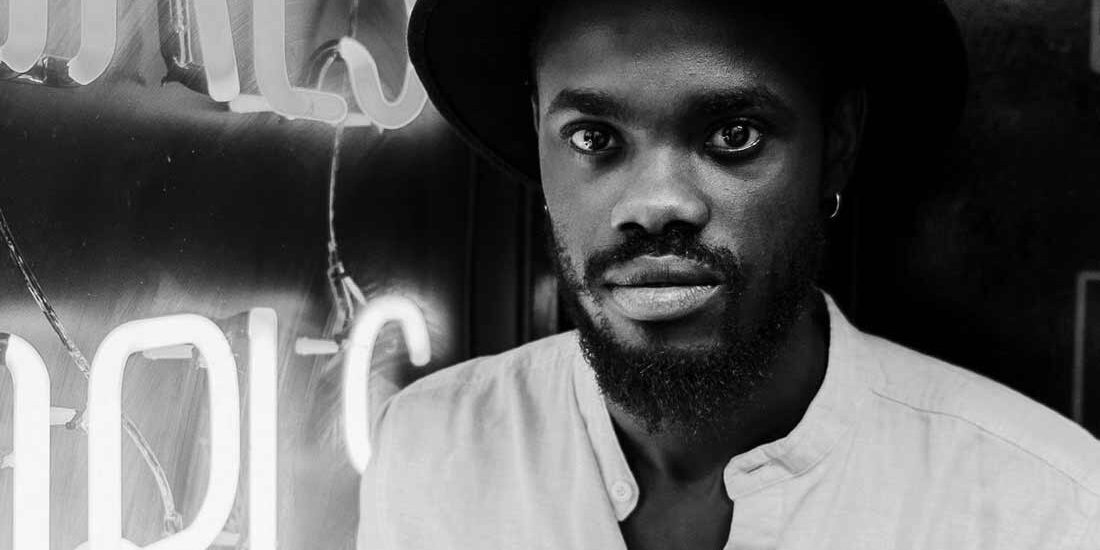 Black and white portrait of a man wearing a hat in front of neon lights.